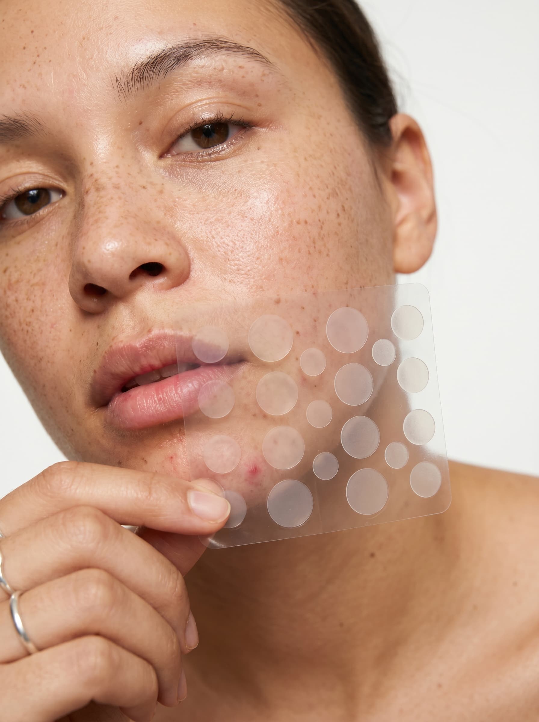 Woman holding a sheet of clear MEND hydrocolloid pimple patches against her cheek, the patches nearly invisible on her skin